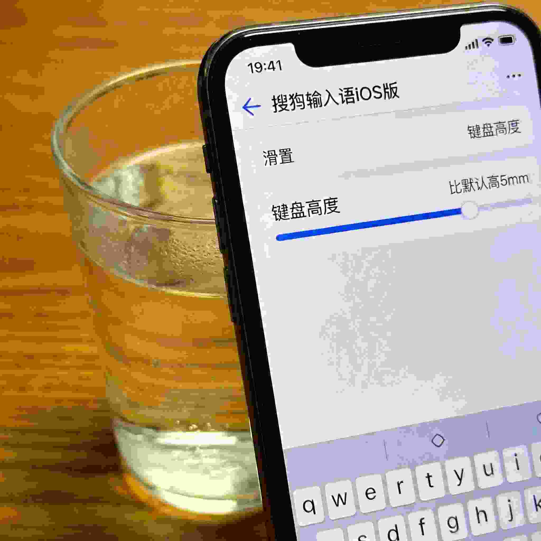 桌面的iPhone屏幕显示搜狗拼音输入法iOS版的键盘设置界面，滑块停在比默认高5mm的位置，旁边放着半杯温水