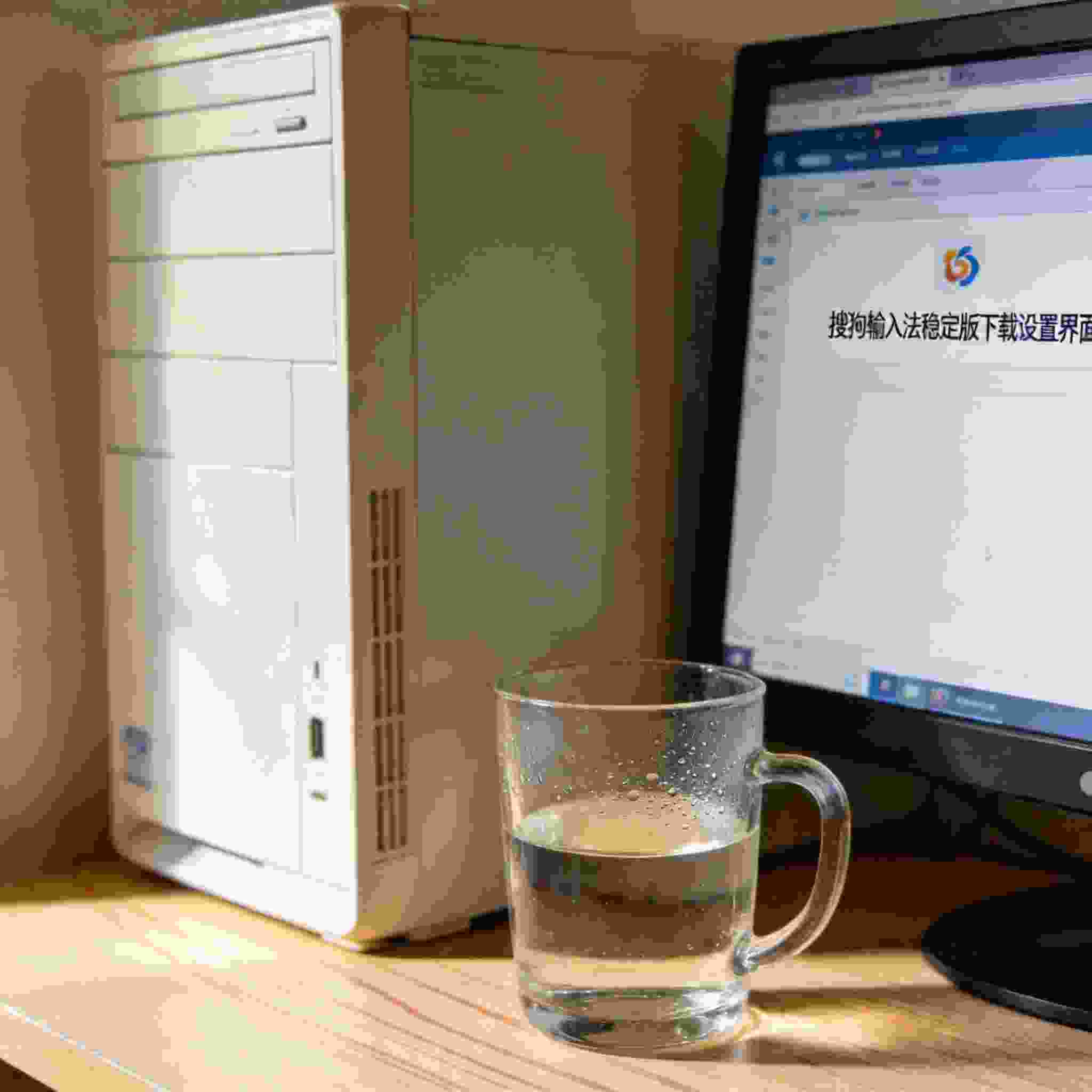 书桌上的主机，屏幕显示搜狗拼音输入法稳定版下载设置界面，桌角放着水杯，自然光
