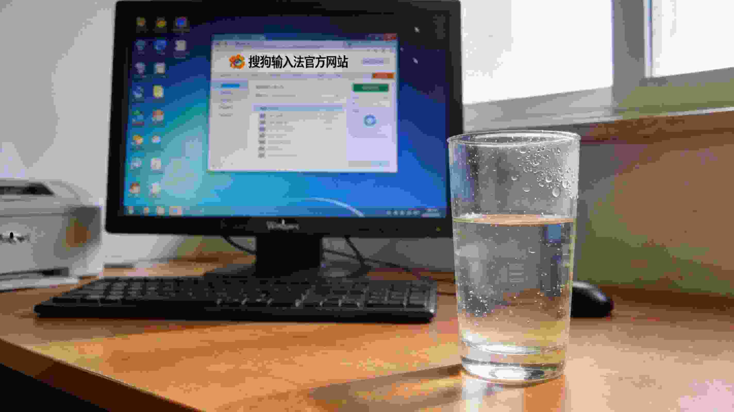电脑桌的Windows系统电脑，屏幕显示搜狗拼音输入法官方网站页面，桌面放着半杯凉白开，室内自然光