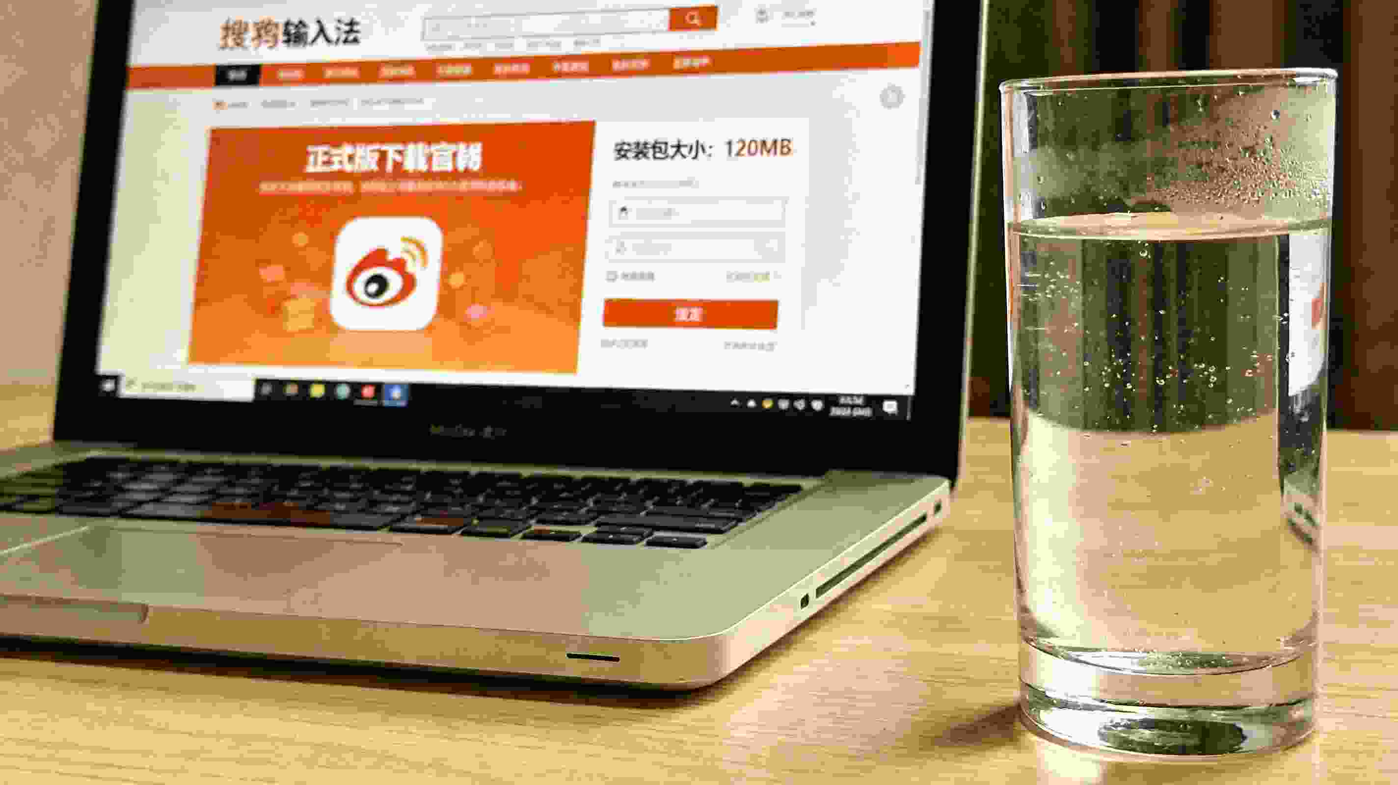 电脑桌面，屏幕显示搜狗拼音输入法官网的正式版下载页面，页面标注安装包大小120MB，旁边放着半杯凉白开，室内自然光