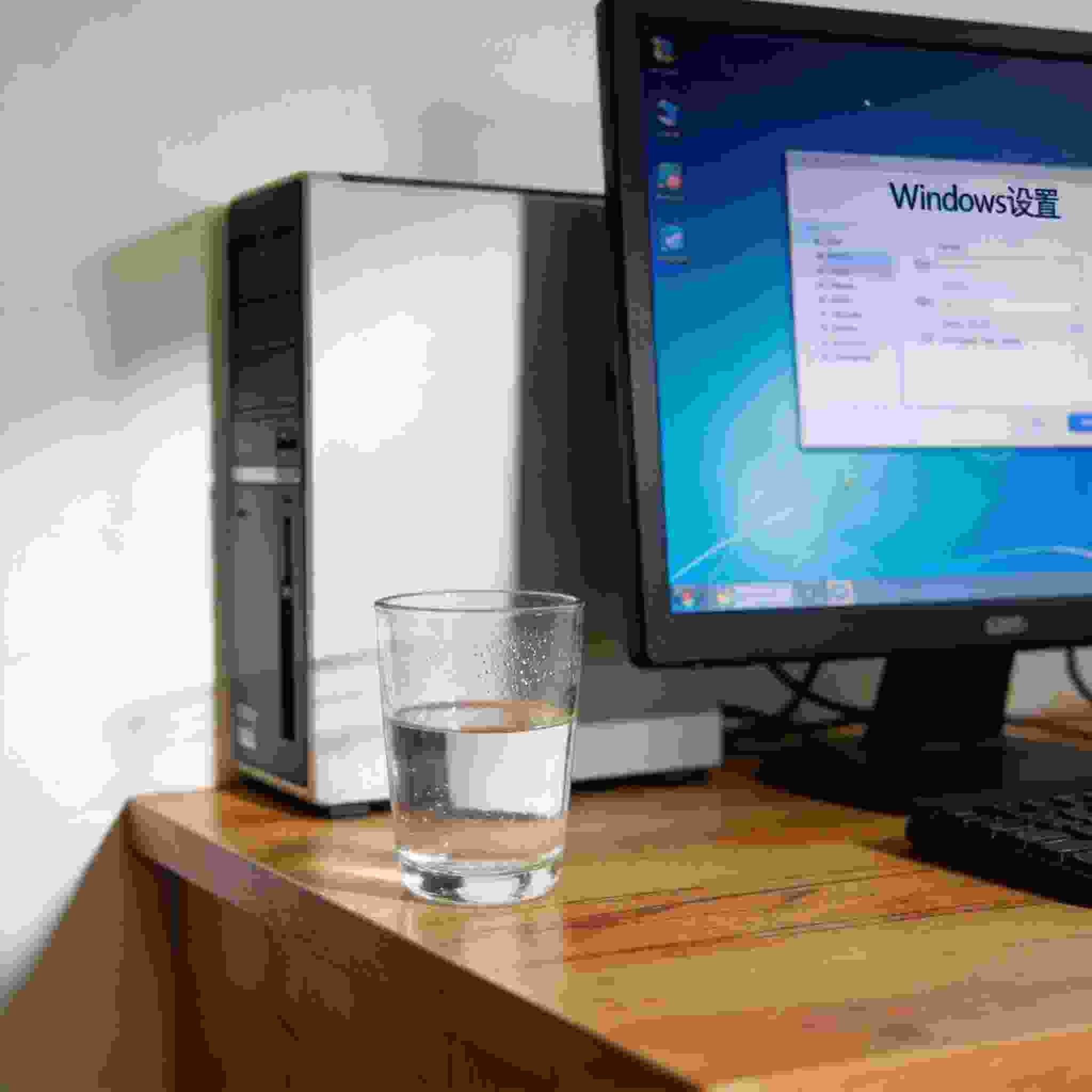 书桌上的主机，屏幕显示Windows设置界面，桌角放着水杯，自然光