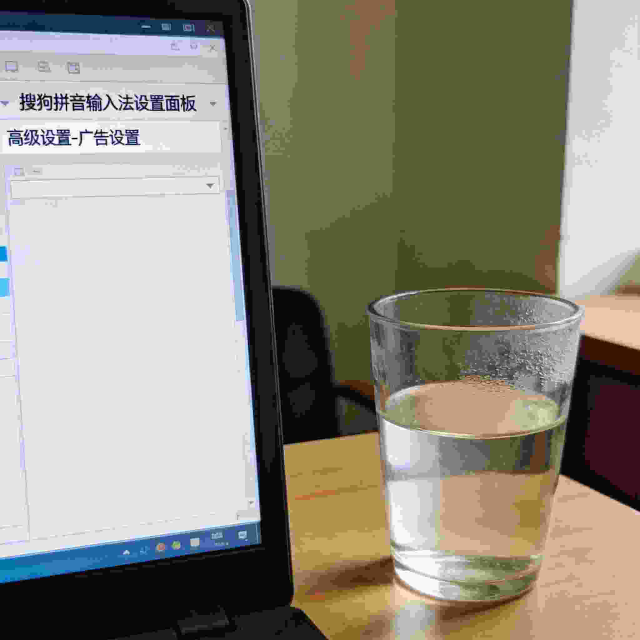 电脑桌的电脑屏幕显示搜狗拼音输入法设置面板，停在高级设置的广告设置页面，桌面放着半杯温水，室内自然光