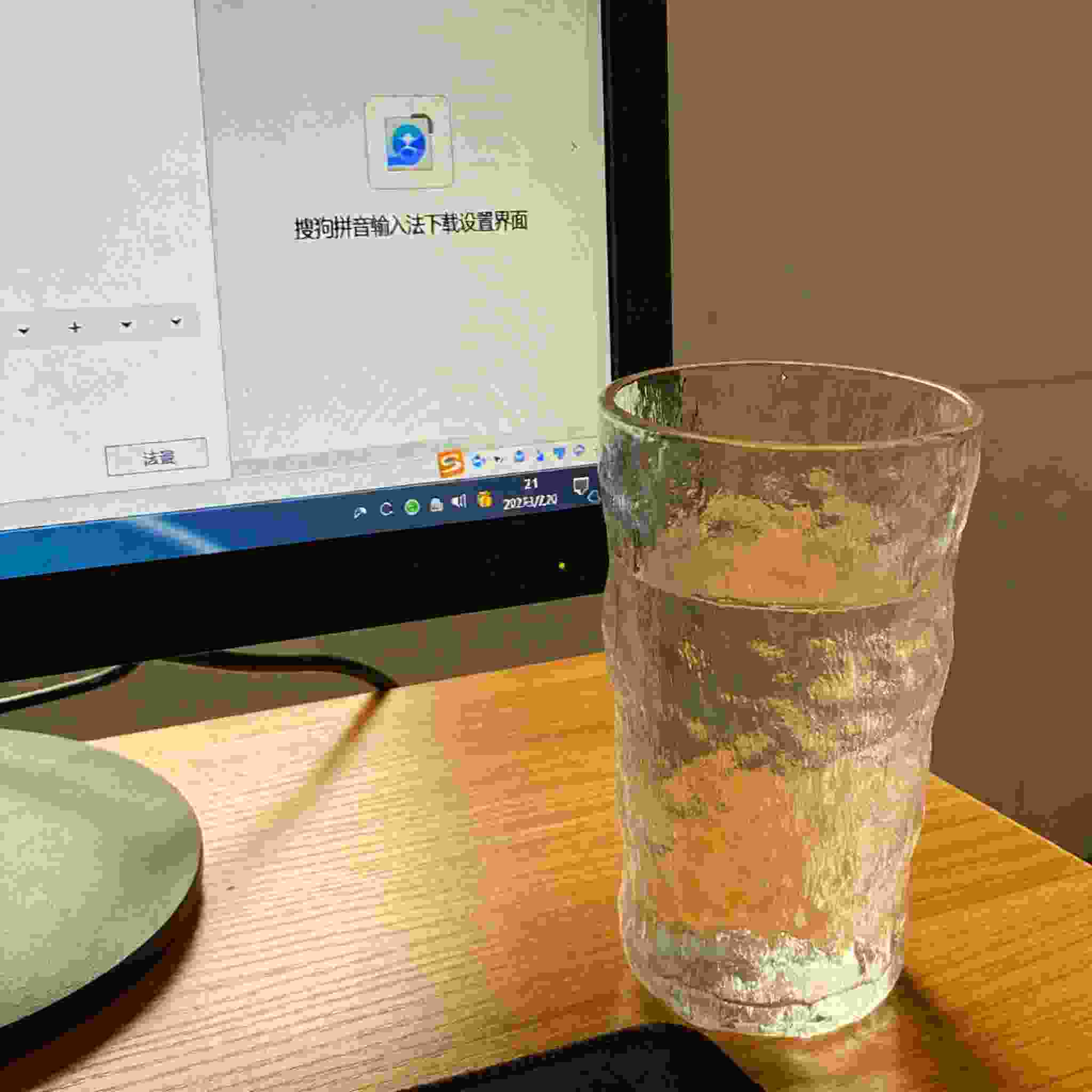 书桌上的主机，屏幕显示搜狗拼音输入法下载设置界面，桌角放着水杯，自然光