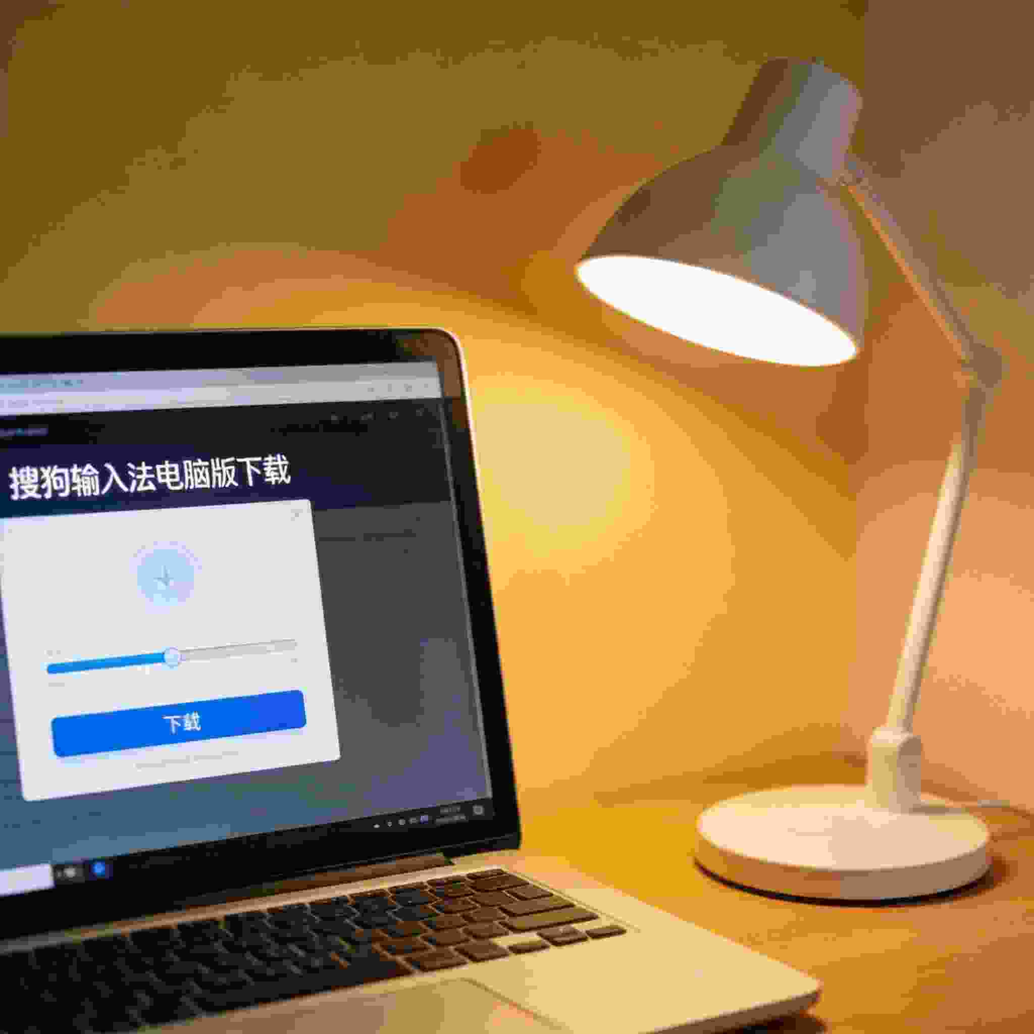 电脑桌上的电脑，屏幕显示搜狗拼音输入法电脑版下载界面，旁边放着台灯