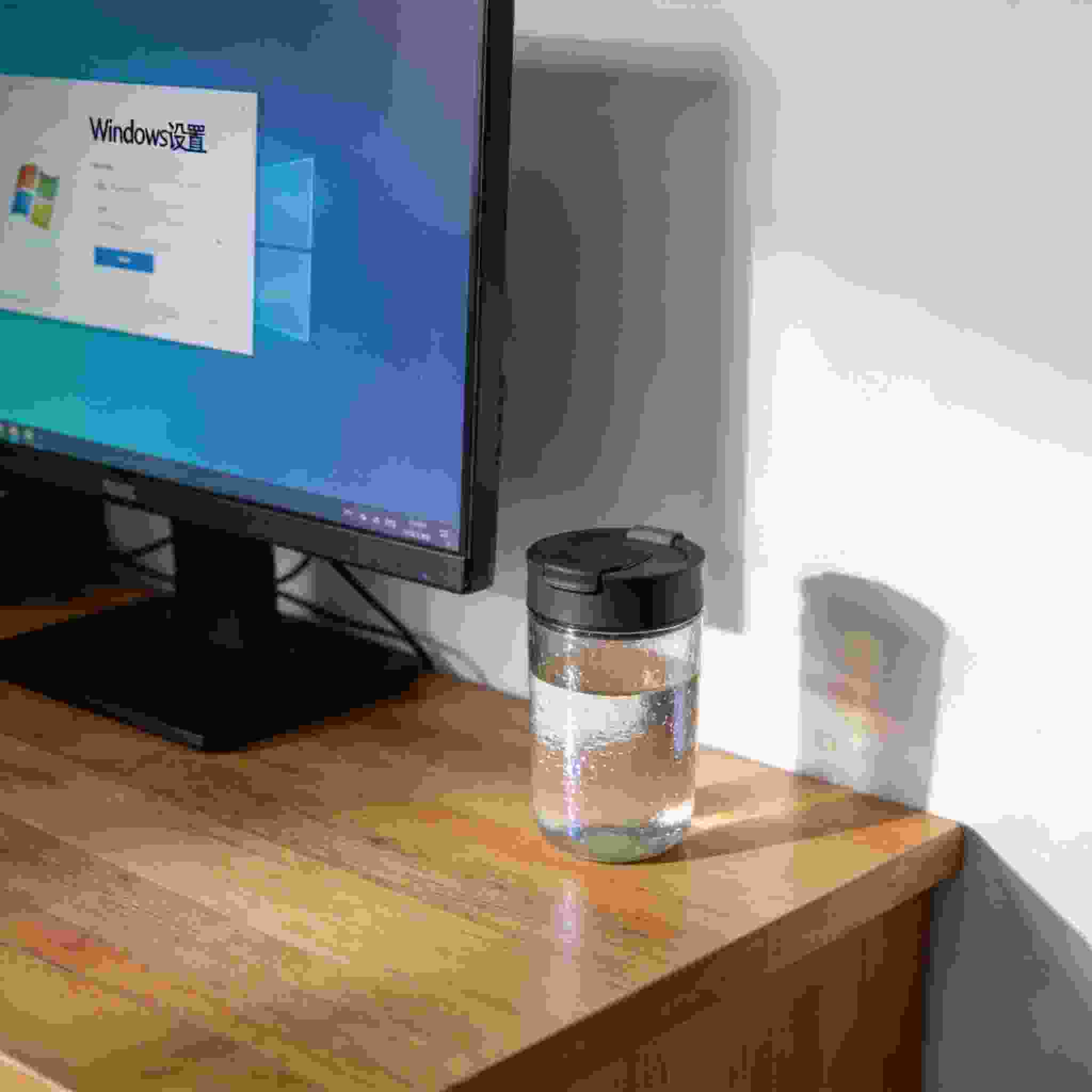 书桌上的主机，屏幕显示Windows设置界面，桌角放着水杯，自然光