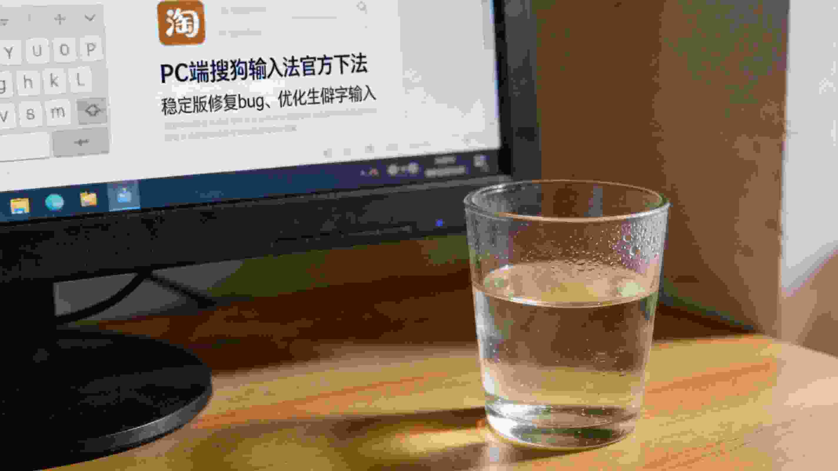 电脑桌面，屏幕显示PC端搜狗拼音输入法官方下载页面，页面标注稳定版修复bug、优化生僻字输入内容，桌面角落放着半杯温水，室内自然光