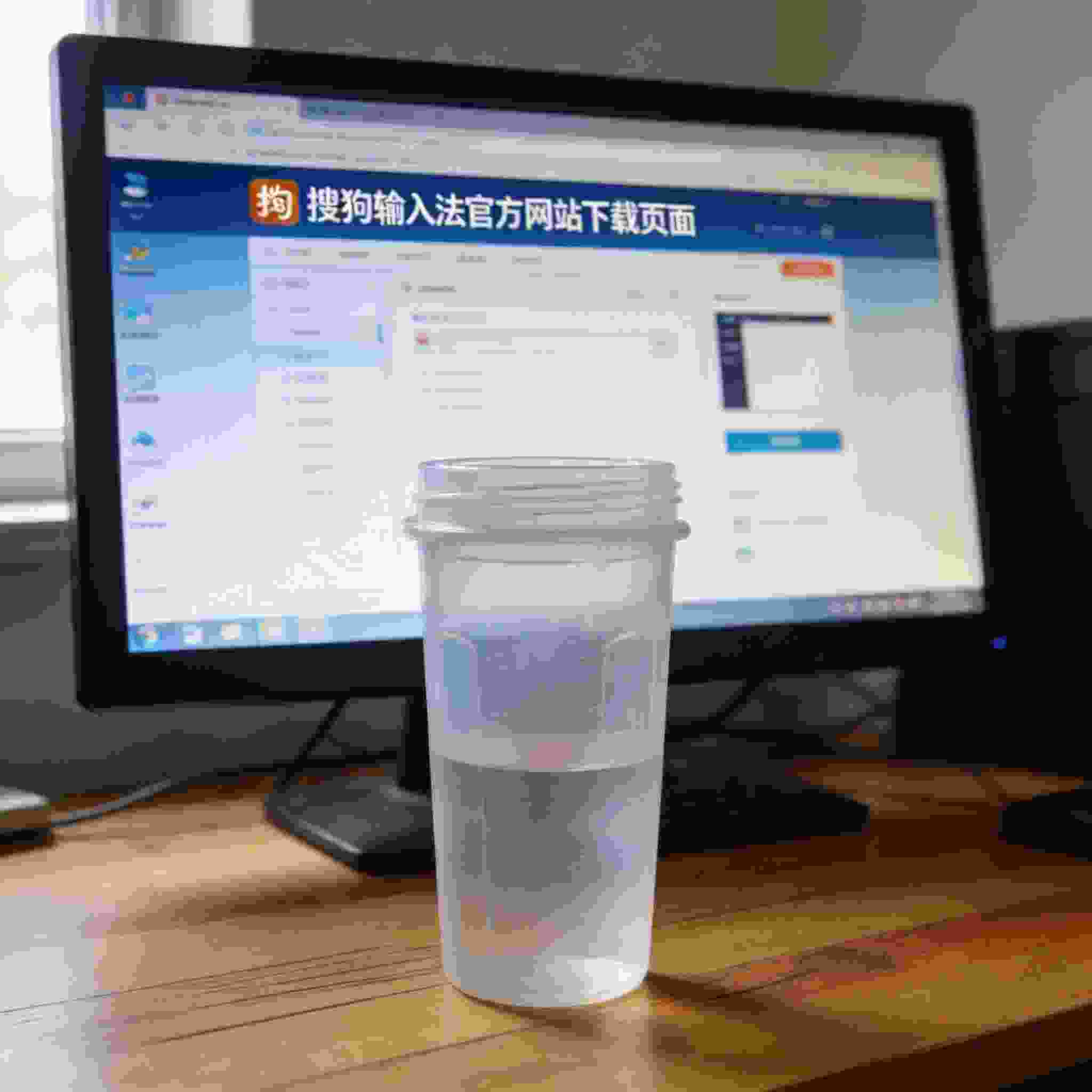 电脑桌的Windows系统电脑，屏幕显示搜狗拼音输入法官方网站下载页面，桌面放着半透明塑料水杯，室内自然光