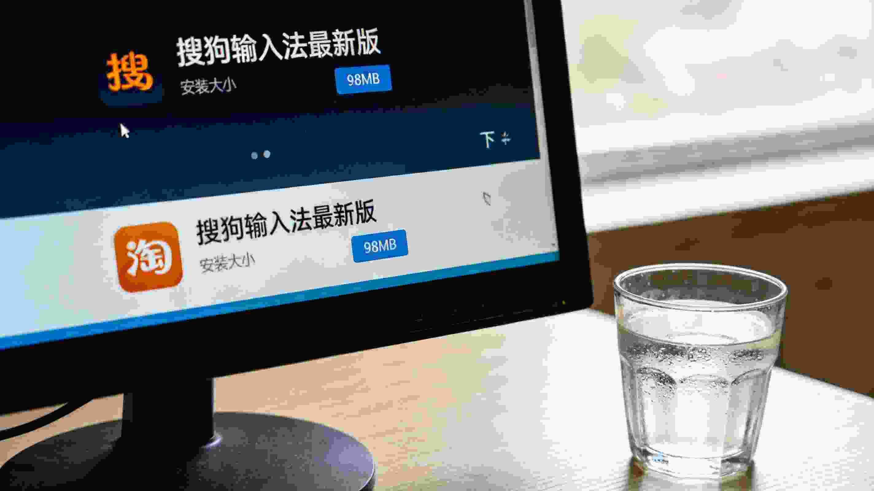 电脑桌面，屏幕显示搜狗拼音输入法最新版下载界面，安装包大小显示98MB，旁边放着半杯凉白开，室内自然光