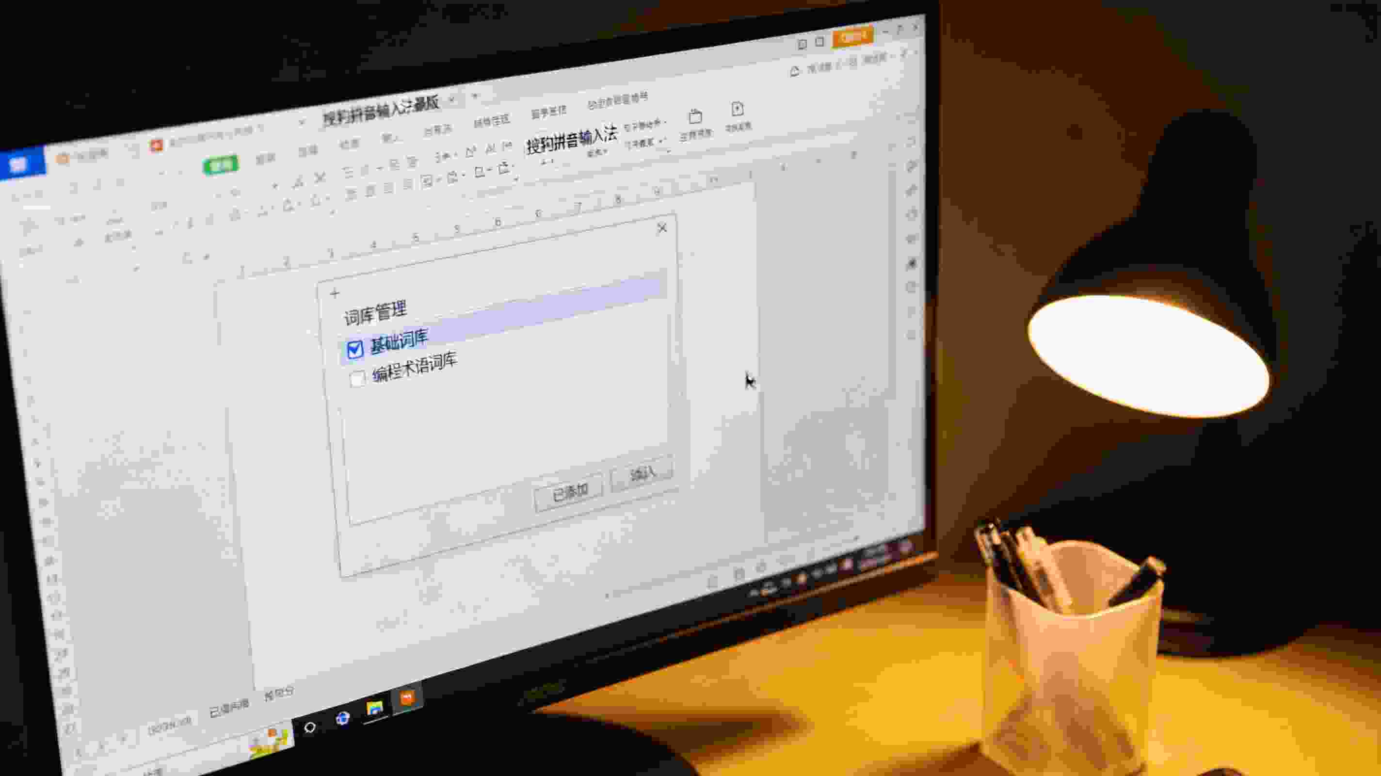 电脑屏幕显示搜狗拼音输入法最新版的词库管理面板，已勾选基础词库，还添加了编程术语词库，桌面角落有个塑料笔筒，台灯暖光