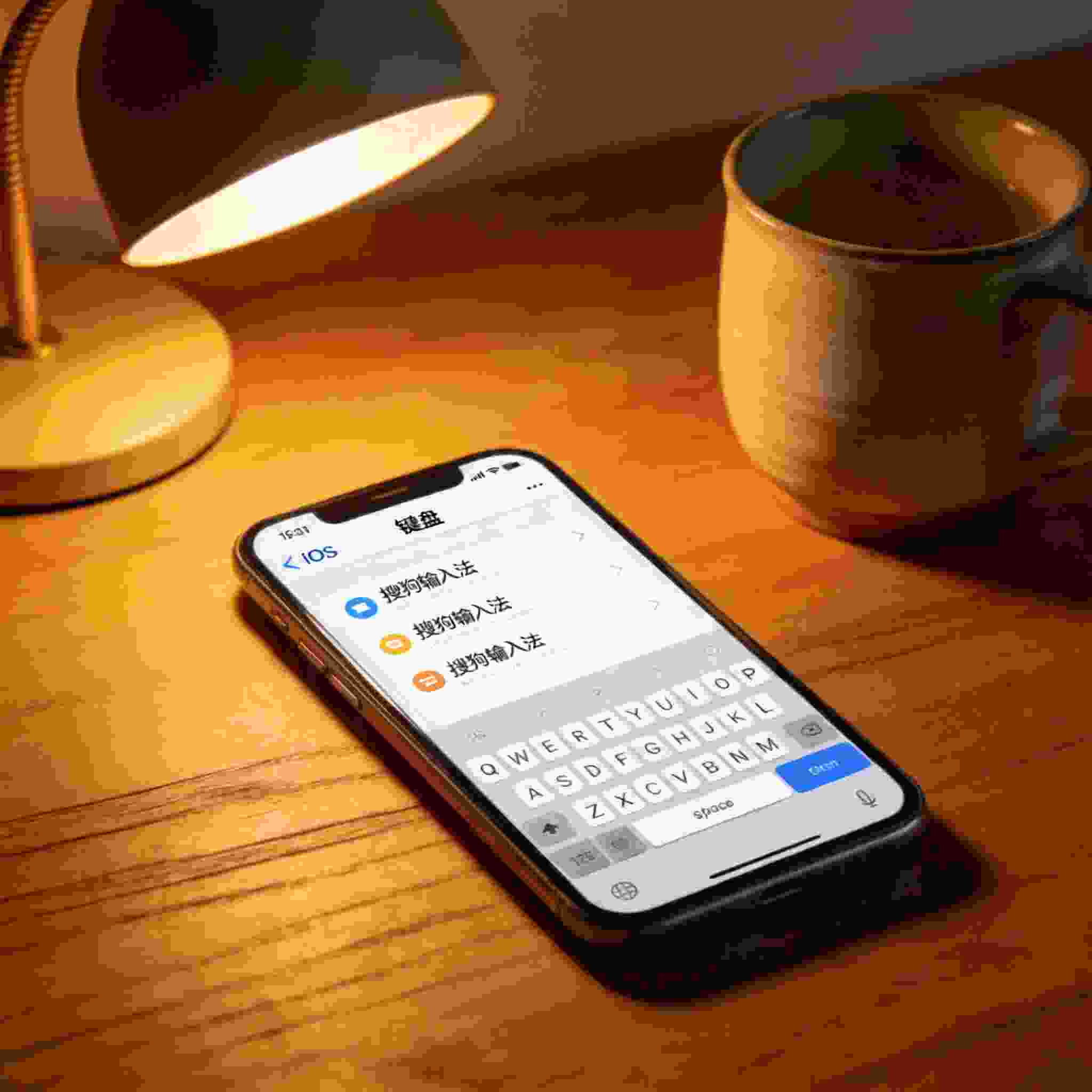桌面的iPhone屏幕显示iOS系统“键盘”设置界面，已添加搜狗拼音输入法，旁边放着陶瓷水杯，台灯暖光