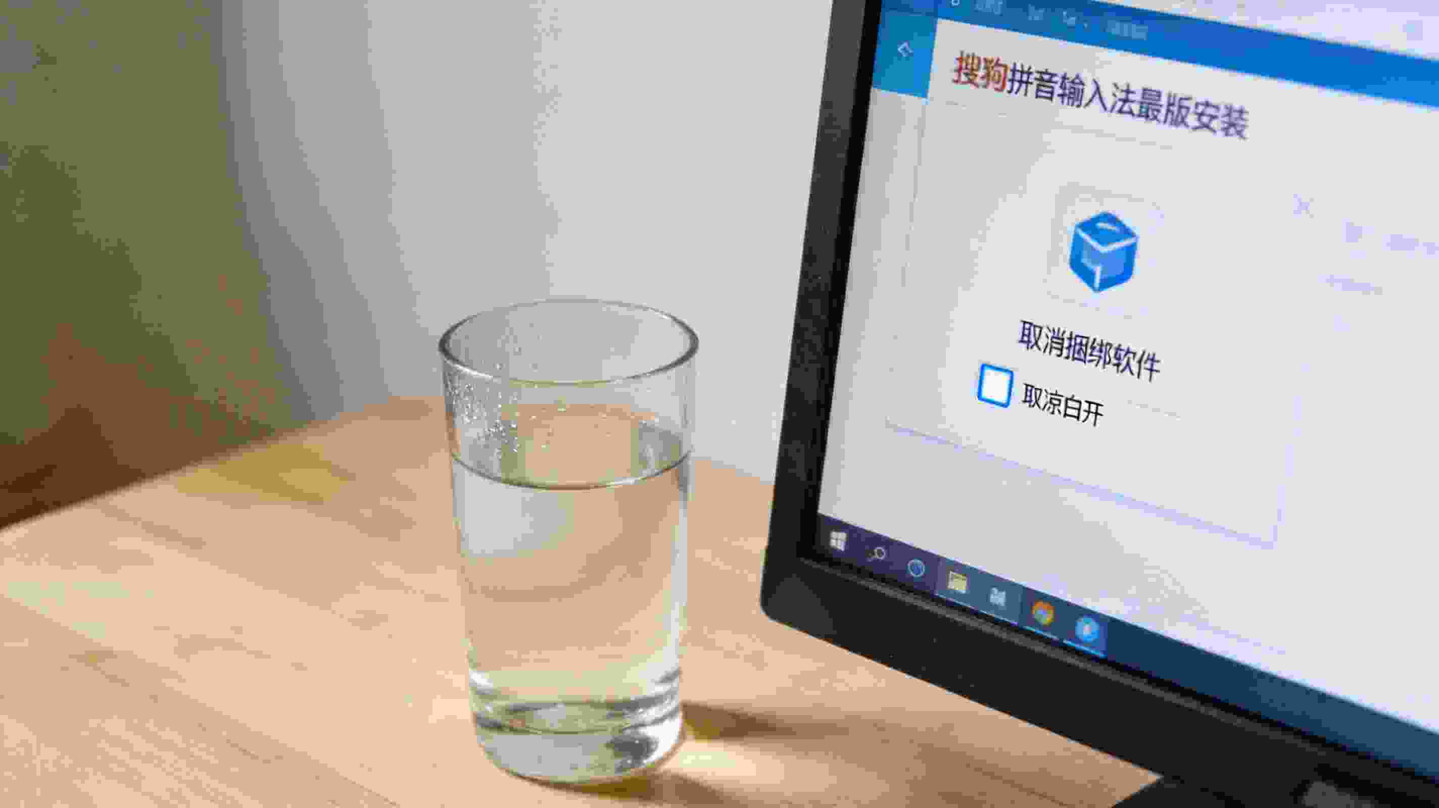 电脑桌面显示搜狗拼音输入法最新版的安装界面，界面上有取消捆绑软件的勾选框，旁边放着半杯凉白开，室内自然光