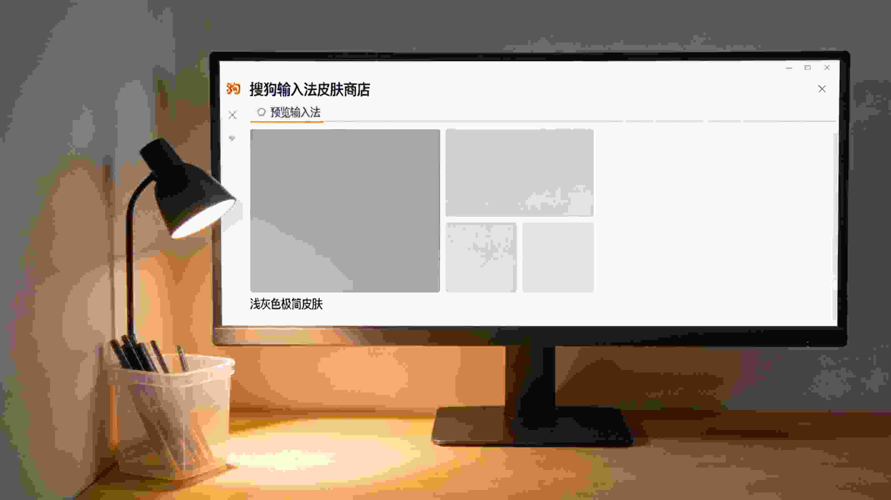 台式机屏幕显示搜狗拼音输入法最新版的皮肤商店界面，浅灰色极简皮肤在预览栏，桌面角落有个塑料笔筒，台灯暖光