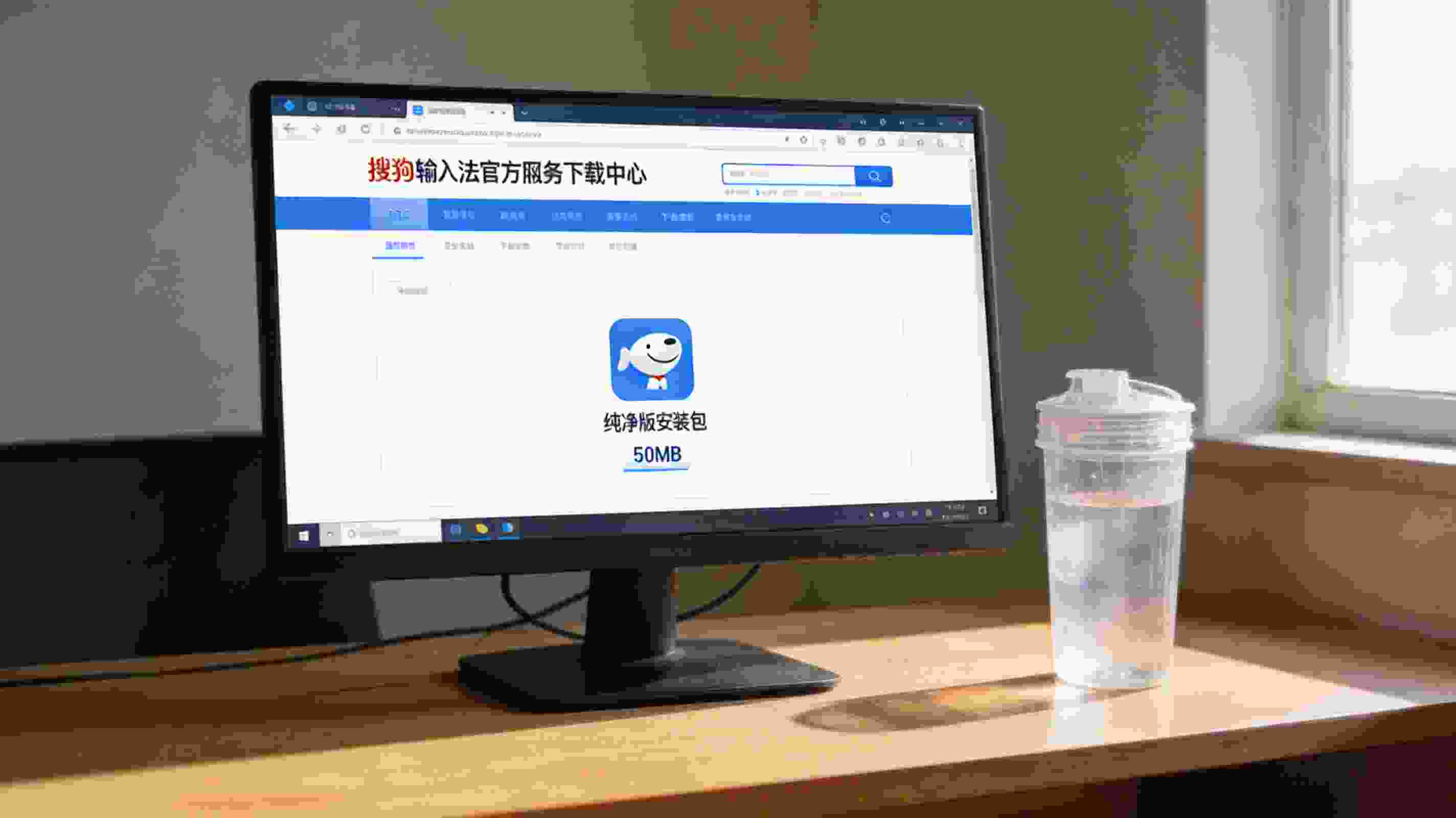 电脑桌面，屏幕显示搜狗拼音输入法官方网站下载中心页面，纯净版安装包标注50MB大小，旁边放着半透明塑料水杯，室内自然光