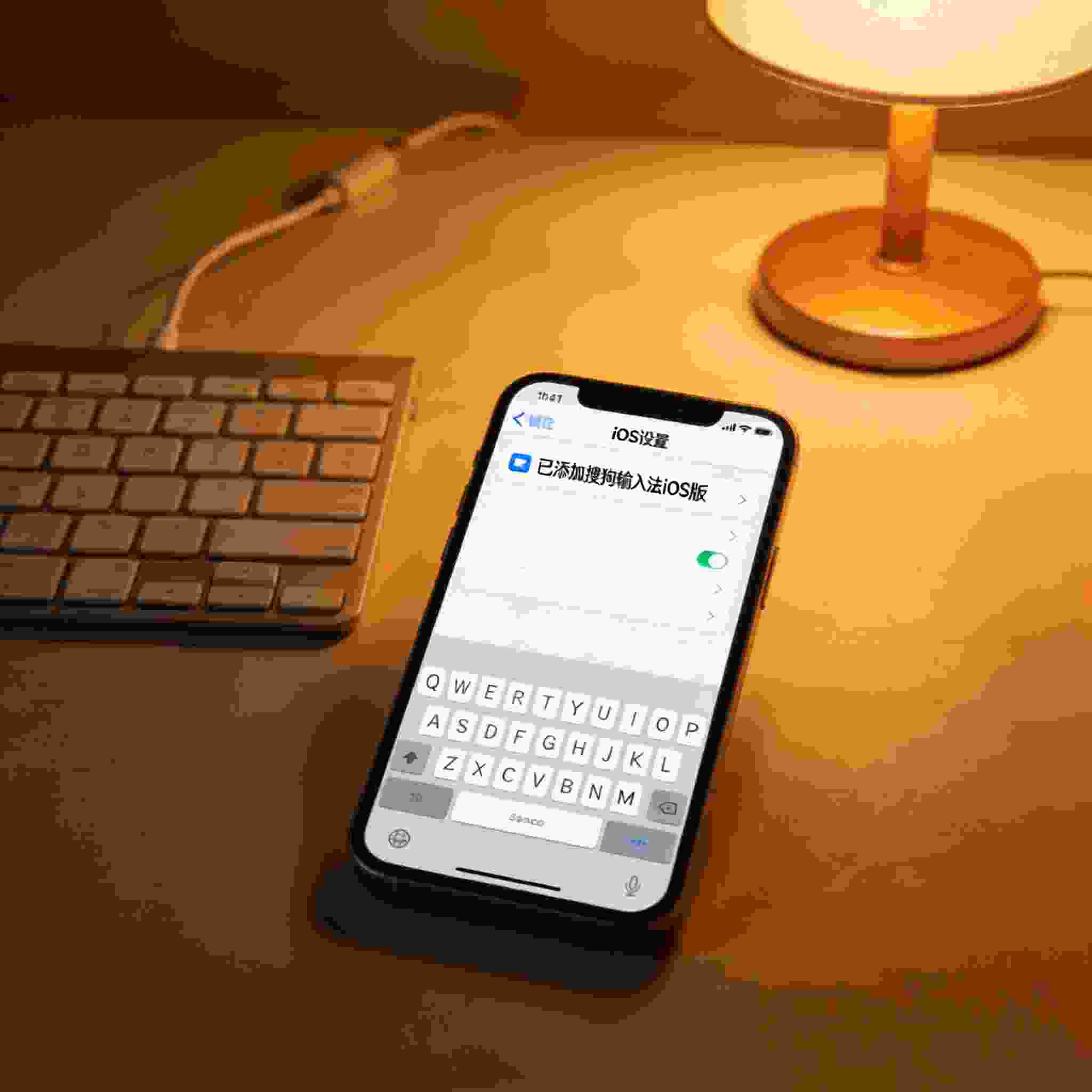 桌面的iPhone屏幕显示iOS系统设置的键盘选项界面，已添加搜狗拼音输入法iOS版，旁边放着有线键盘，台灯暖光