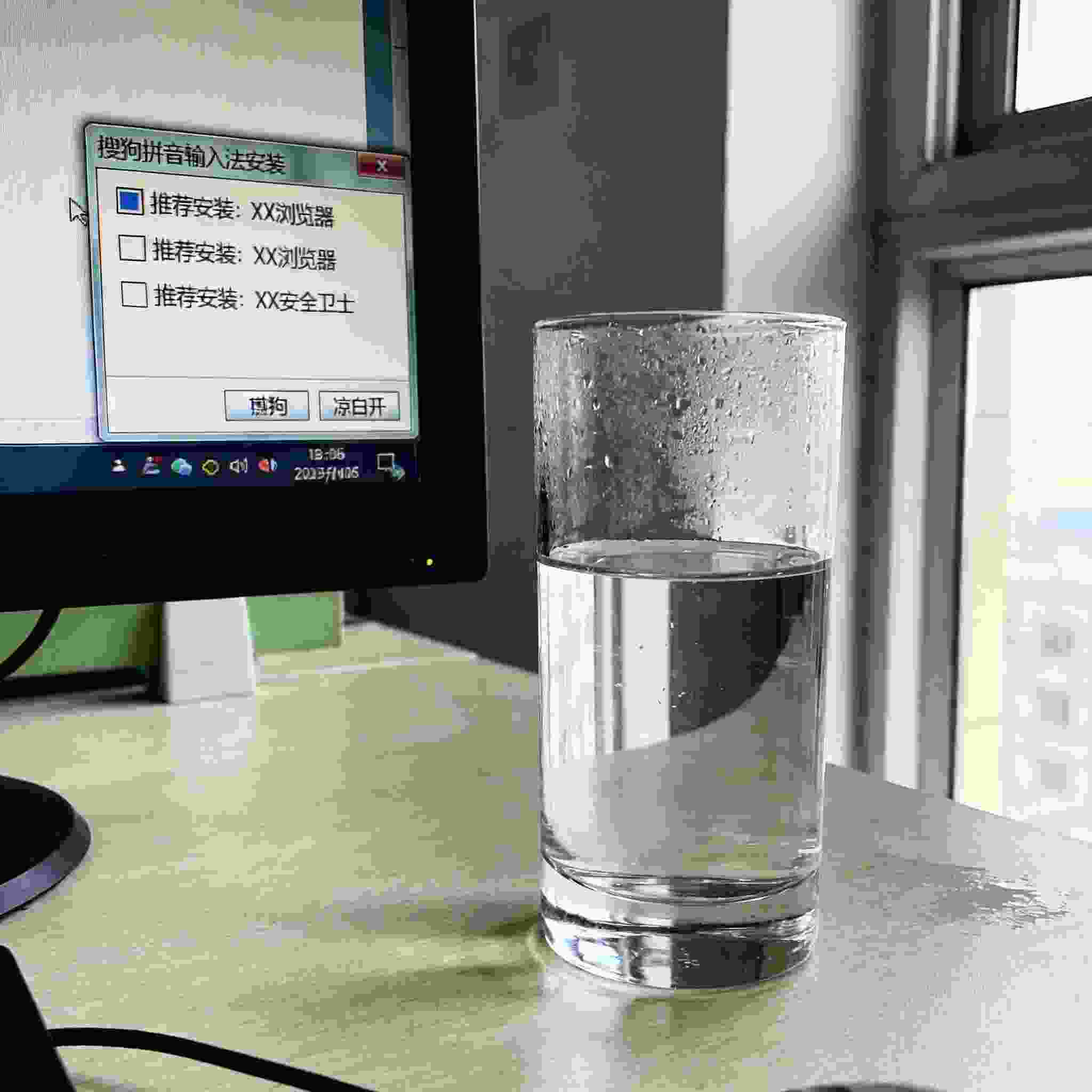 电脑桌的电脑屏幕显示搜狗拼音安装界面，界面上有推荐安装软件的勾选框，桌面放着半杯凉白开，室内自然光