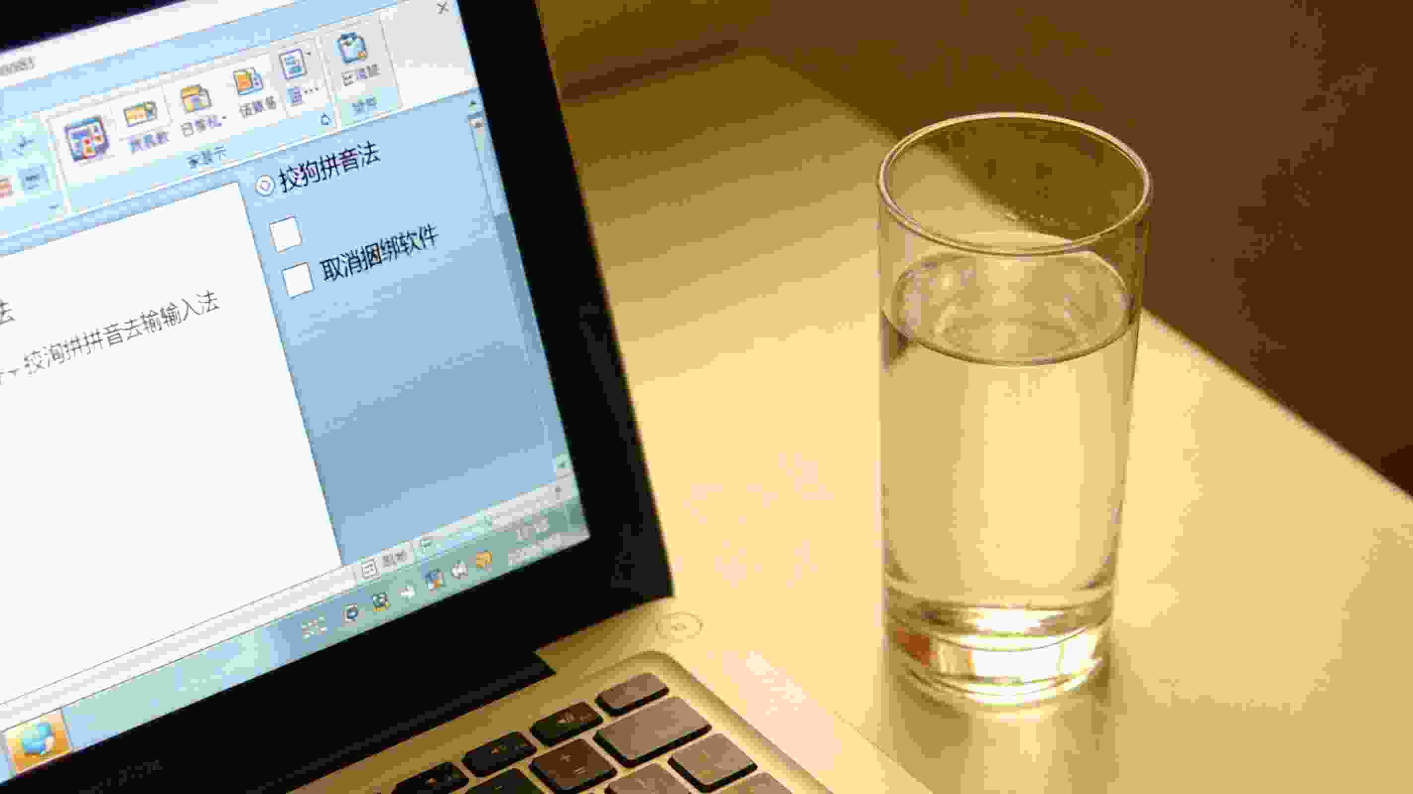 电脑桌面显示搜狗拼音输入法安装界面，界面上有取消捆绑软件的勾选框，旁边放着半杯温水，室内自然光