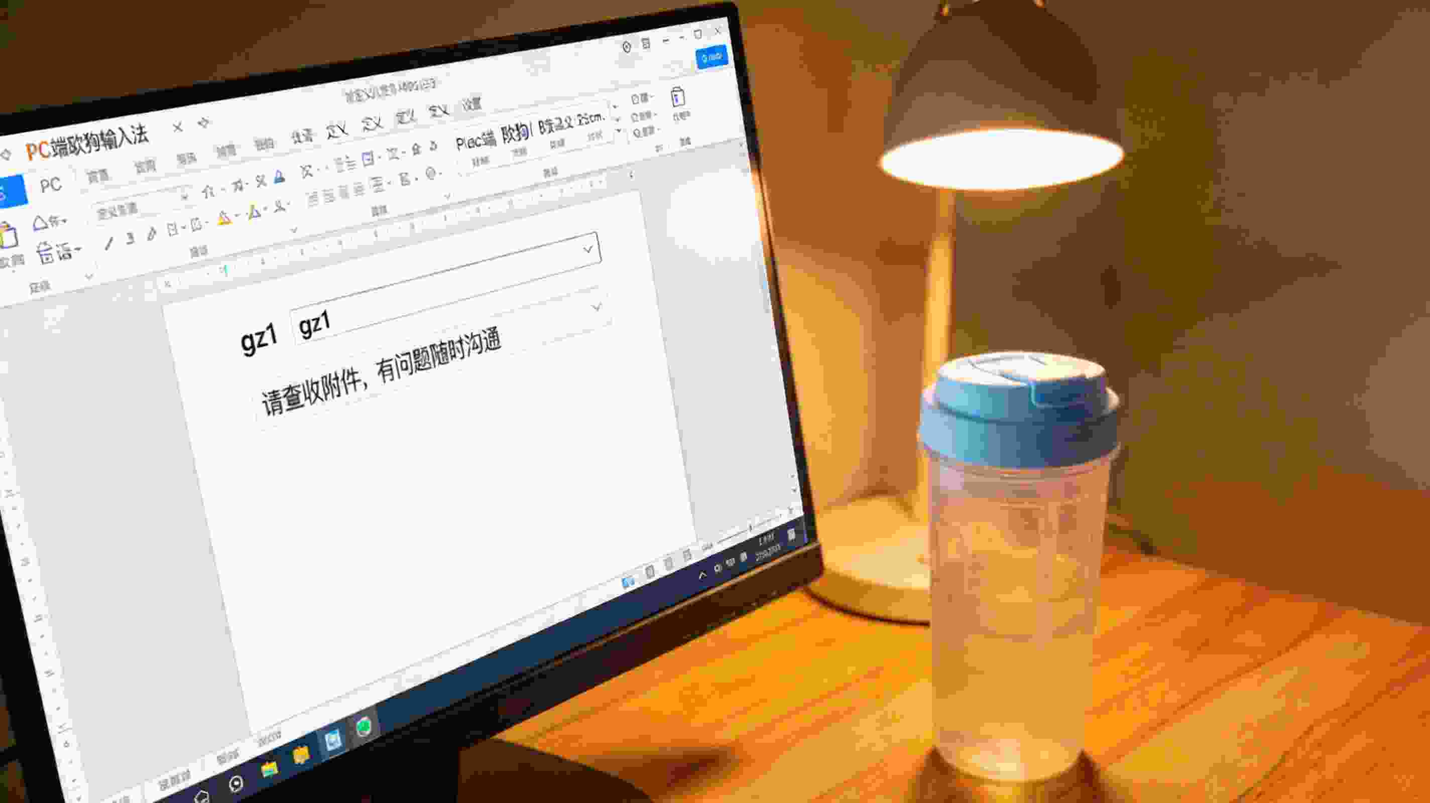 电脑屏幕显示PC端搜狗拼音输入法自定义短语设置界面，输入框内显示“gz1”，对应内容为“请查收附件，有问题随时沟通”，旁边放着普通塑料水杯，台灯暖光