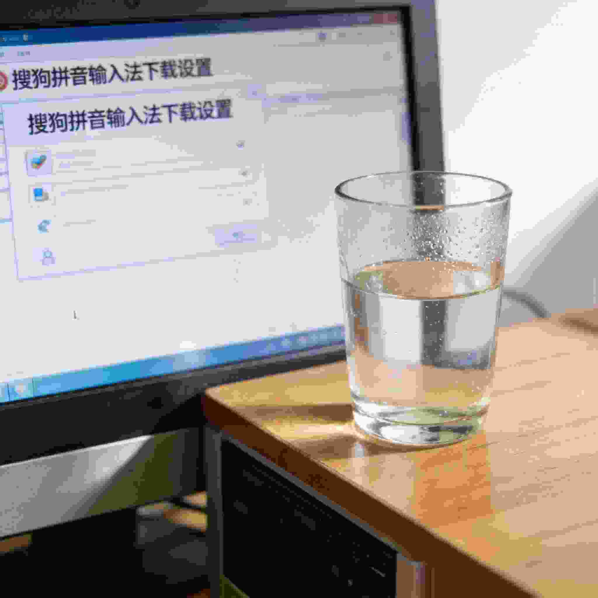 书桌上的主机，屏幕显示搜狗拼音下载设置界面，桌角放着水杯，自然光